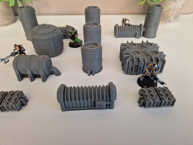 May include: A collection of gray 3D printed miniature buildings and figures. The buildings are various shapes and sizes, including cylinders, rectangular boxes, and a square building with four round vents. There are also two miniature figures, one green and one gold.