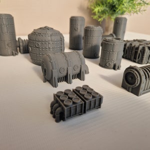 May include: A collection of gray, 3D printed, industrial-style miniature buildings. The buildings are made of various shapes and sizes, including cylinders, cubes, and rectangular structures. Some of the buildings have intricate details, such as pipes and vents.