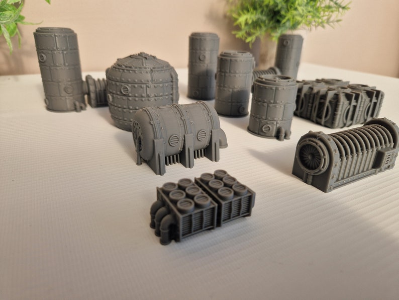 May include: A collection of gray, 3D printed, industrial-style miniature buildings. The buildings are made of various shapes and sizes, including cylinders, cubes, and rectangular structures. Some of the buildings have intricate details, such as pipes and vents.