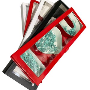May include: A set of four gift boxes with clear windows, each box is a different color: red, black, white, and a lighter shade of red. The boxes are stacked on top of each other. The boxes are filled with teal and white shredded paper.