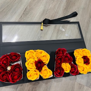 May include: A black box with a clear window, featuring the word "GRAD" spelled out with red and yellow roses. A black ribbon and gold tassel are attached to the box.
