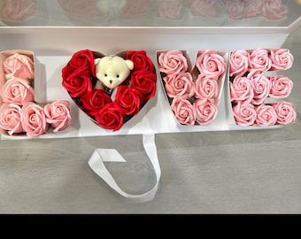 May include: A white box with the word "LOVE" spelled out in pink and red roses. The letter "O" is a heart-shaped box with red roses and a small teddy bear in the center.