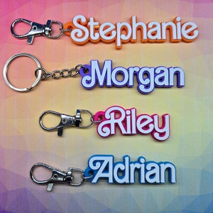 May include: Four colourful keychains with the names "Stephanie", "Morgan", "Riley", and "Adrian" written in white letters. The keychains are made of plastic and have a silver metal clasp.