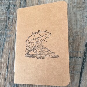 Peut inclure: Un carnet en papier brun avec une illustration en noir et blanc d'une grenouille assise sous un parapluie sous la pluie. La grenouille tient le parapluie au-dessus de sa tête. Des gouttes de pluie tombent du parapluie.