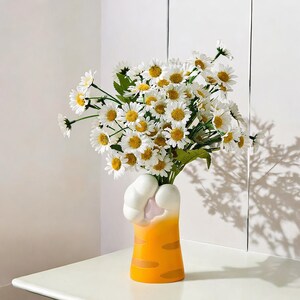 May include: A white ceramic vase shaped like a cat paw with orange and white stripes holds a bouquet of white daisies.