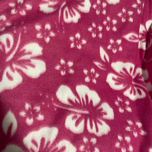 Op de afbeelding: Een zachte stof met een levendige magentakleurige achtergrond en een herhalend patroon van grote en kleine witte hibiscusbloemen. Het materiaal heeft een pluizige, zachte textuur en toont een consistent bloemenpatroon over het oppervlak.