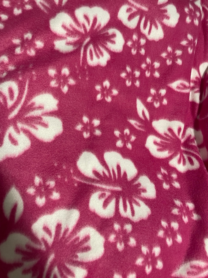 Op de afbeelding: Een zachte stof met een levendige magentakleurige achtergrond en een herhalend patroon van grote en kleine witte hibiscusbloemen. Het materiaal heeft een pluizige, zachte textuur en toont een consistent bloemenpatroon over het oppervlak.