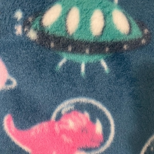 Op de afbeelding: Een zachte blauwe fleecestof met een herhalend patroon van groenblauwe ufo's met witte accenten en roze dinosaurussen die doorzichtige ruimtehelmen dragen. Het materiaal heeft een pluizige textuur en een print met een ruimtethema.