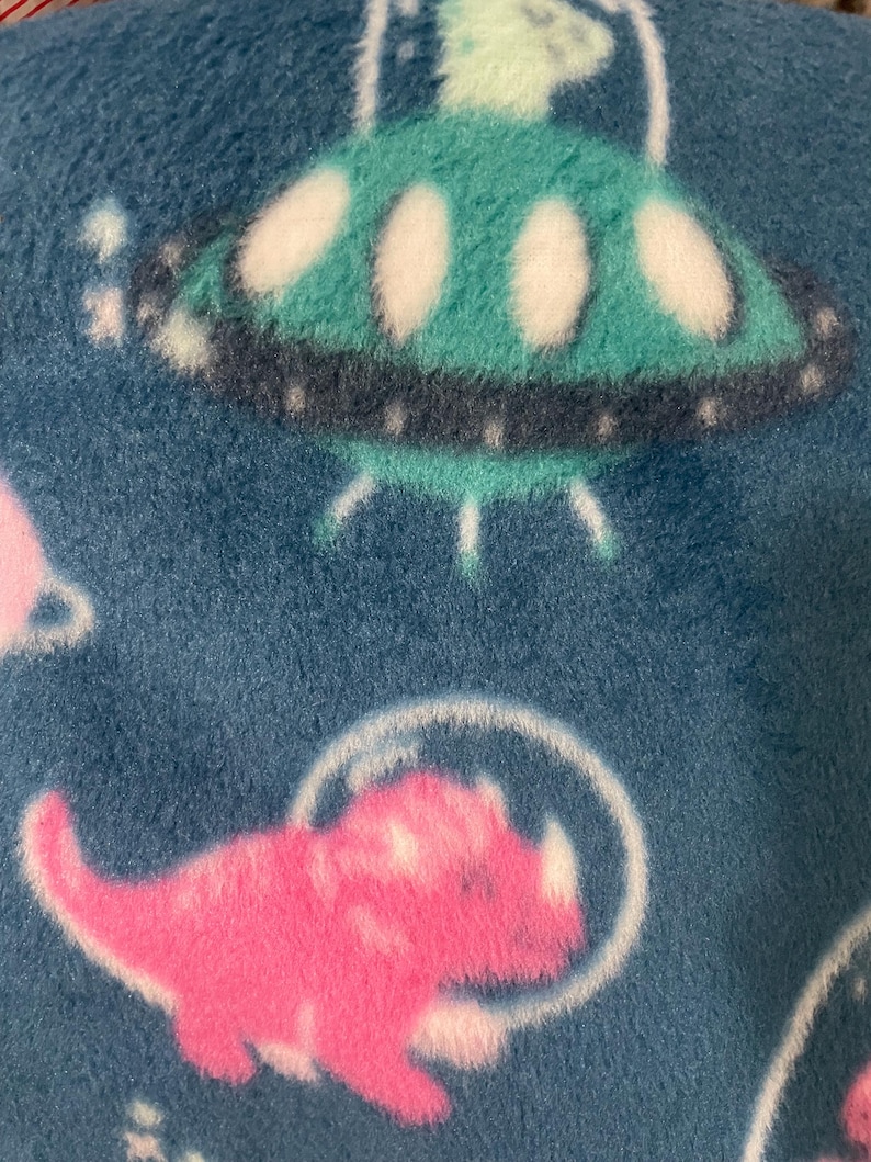 Op de afbeelding: Een zachte blauwe fleecestof met een herhalend patroon van groenblauwe ufo's met witte accenten en roze dinosaurussen die doorzichtige ruimtehelmen dragen. Het materiaal heeft een pluizige textuur en een print met een ruimtethema.