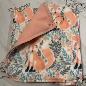 May include: A reversible pet bed with a fox and floral pattern. The outer layer features orange foxes and botanical elements on a white background. The inner layer is a solid peach color. Metal clips are attached to the corners.