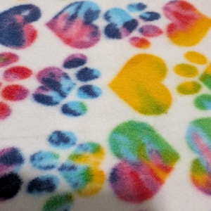 Op de afbeelding: Een witte stof met een herhalend patroon van tie-dye harten en pootafdrukken in levendige kleuren, waaronder blauw, roze, geel en groen. Het zacht aanvoelende materiaal vertoont een veelkleurig aquareleffect op elke vorm.