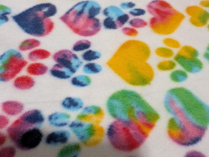 Op de afbeelding: Een witte stof met een herhalend patroon van tie-dye harten en pootafdrukken in levendige kleuren, waaronder blauw, roze, geel en groen. Het zacht aanvoelende materiaal vertoont een veelkleurig aquareleffect op elke vorm.