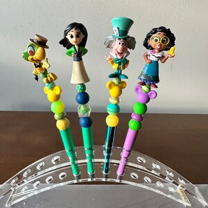 May include: Four colorful Disney character themed pens with beads and charms on a clear acrylic pen holder. The characters are Mulan, the Mad Hatter, Tiana, and a green bird.