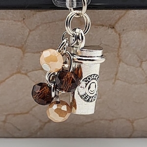 May include: Silver-colored coffee cup charm with the word "Coffee" and a smiling face. The charm is attached to a silver ring and has a cluster of brown and tan beads. The background is a textured, neutral color.