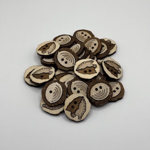 May include: A pile of wooden buttons with a brown and white design. The buttons feature a stylized image of a Native American arrowhead.