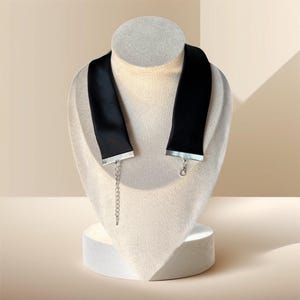 May include: A black choker necklace displayed on a mannequin. The choker is made of a wide, black satin ribbon with silver-colored trim at the ends. A silver chain extends from one end for adjustable sizing. The necklace is a fashion accessory.
