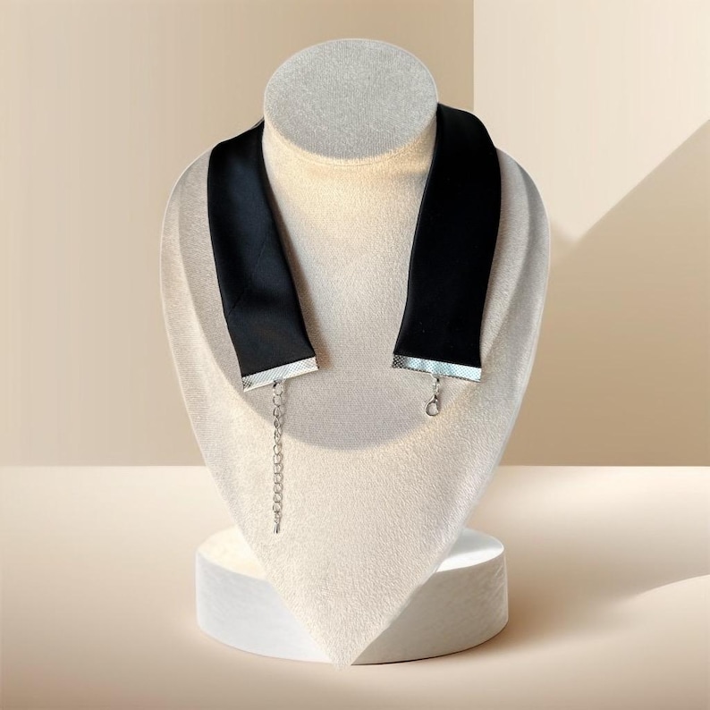May include: A black choker necklace displayed on a mannequin. The choker is made of a wide, black satin ribbon with silver-colored trim at the ends. A silver chain extends from one end for adjustable sizing. The necklace is a fashion accessory.