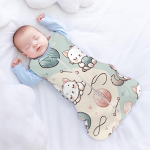 Può includere: Un bambino dorme in un sacco a pelo blu chiaro e verde con un motivo di gattini bianchi, gomitoli di lana e spilli.