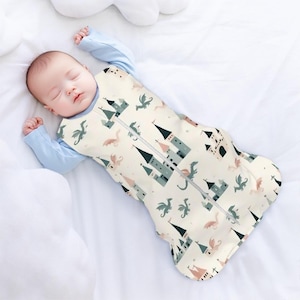 Op de afbeelding: Een witte baby slaapzak met een ritssluiting. De slaapzak heeft een patroon van groene kastelen en draken op een crème achtergrond.