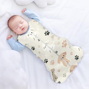 May include: A cream-colored baby sleeping bag with a zipper closure. The sleeping bag features a pattern of brown and white dogs and paw prints.