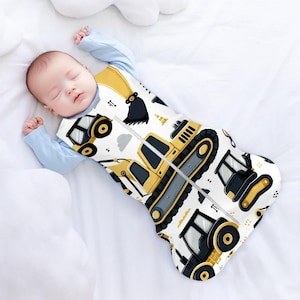 Puede incluir: Un saco de dormir para bebé blanco y amarillo con cierre de cremallera. El saco de dormir presenta un patrón de vehículos de construcción, incluyendo excavadoras, bulldozers y camiones de volteo.