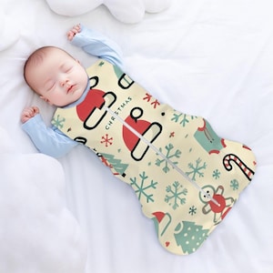 May include: A baby sleeping in a cream-coloured, Christmas-themed swaddle sack with a zipper. The sack has a red and white Santa hat, a green Christmas tree, snowflakes, and the word "Christmas" on it.