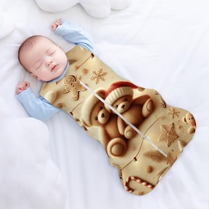 May include: A beige and brown baby sleeping bag with a Christmas theme. The sleeping bag features two teddy bears wearing Santa hats, a gingerbread man, and snowflakes. The sleeping bag has a zipper down the front.