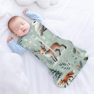 May include: A baby sleeping in a green and white sleeping bag with a deer, owl, and fox print. The sleeping bag has a zip down the front.