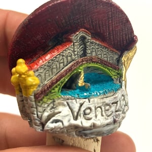 May include: A colorful, detailed, miniature replica of a bridge in Venice, Italy. The bridge is made of a light gray material with red, green, and yellow accents. The bridge is mounted on a cork base and has the word "Venezia" written on the side.