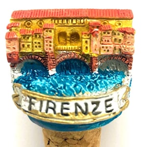 May include: A colorful bottle stopper featuring a miniature replica of the Ponte Vecchio bridge in Florence, Italy. The bridge is painted in shades of red, yellow, and orange, with a blue river flowing beneath it. The stopper is topped with a cork base and a banner that reads "FIRENZE".