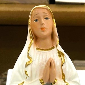 May include: A white figurine of the Virgin Mary, wearing a blue robe and a gold-trimmed white cloak. She is standing on a gold base with red flowers.