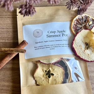 May include: A brown paper bag with a clear plastic window showing dried apple, lemon, and cinnamon slices. The bag has a white label with the text "Crisp Apple Simmer Pot" and the ingredients: "Apple, Lemon, Cinnamon, Cloves".