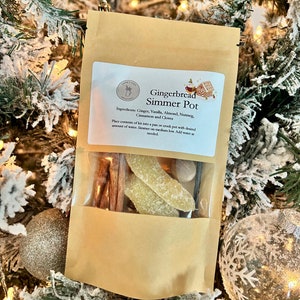 May include: A brown paper bag with a clear window showing the contents of a gingerbread simmer pot kit. The kit includes cinnamon sticks, cloves, vanilla beans, and candied ginger. The bag has a label that reads "Gingerbread Simmer Pot" and lists the ingredients: Ginger, Vanilla, Almond, Nutmeg, Cinnamon and Cloves. The label also includes instructions for using the kit.