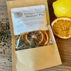 May include: A brown paper bag with a white label that reads "Energy + Focus Simmer Pot" and lists the ingredients: Orange, Lemon, Rosemary, Peppermint. The bag contains dried orange slices, rosemary, and other herbs.