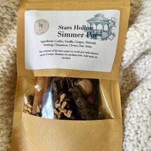 May include: A brown paper bag with a white label that says "Stars Hollow Simmer Pot". The label also lists the ingredients: Coffee, Vanilla, Ginger, Almond, Nutmeg, Cinnamon, Cloves, Star Anise. The bag contains a clear plastic pouch with a mix of spices and coffee beans.