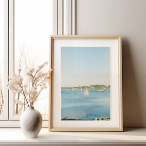 Puede incluir: Una fotografía enmarcada de un paisaje marítimo con veleros en un cuerpo de agua azul tranquilo. El cielo es de un azul claro con nubes blancas. La foto está enmarcada en un marco de madera claro.