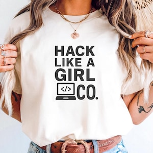 May include: Ivory-colored t-shirt with the text "HACK LIKE A GIRL CO." in black. The design includes a laptop icon with code symbols. The shirt is paired with light blue denim shorts and a brown belt.