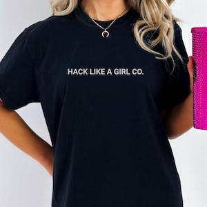 Może przedstawiać: Czarny t-shirt z białym napisem "HACK LIKE A GIRL CO.". Osoba trzyma różowy kubek z ćwiekami i słomką. Koszulka ma wygodny, obszerny krój.