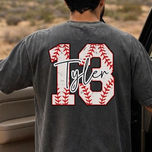 Könnte beinhalten: Ein dunkelgraues T-Shirt mit Baseball-Design. Die Zahl "18" ist in einem Baseball-Nahtmuster dargestellt, mit dem Namen "Tyler" in Schreibschrift. Das T-Shirt hat kurze Ärmel.