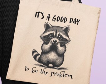 May include: A cream tote bag with a black and white illustration of a raccoon. The raccoon is depicted in a seated pose, with its paws near its face. The text "IT'S A GOOD DAY to be the problem" is printed above and below the raccoon.