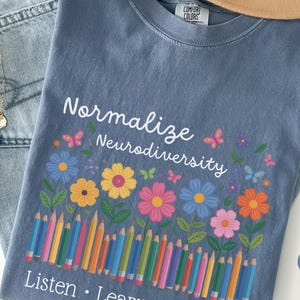 Può includere: Una maglietta blu jeans con le parole "Normalize Neurodiversity" in bianco, insieme a fiori colorati, farfalle e matite. Un cappello color cachi e jeans azzurri sono anche nell'immagine.