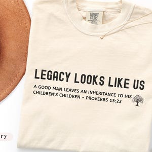 Könnte beinhalten: Elfenbeinfarbenes T-Shirt mit dem Text "LEGACY LOOKS LIKE US" und einem Bibelvers. Ein brauner Hut und eine goldene Halskette sind sichtbar. Das Shirt hat ein Comfort Colors-Etikett.