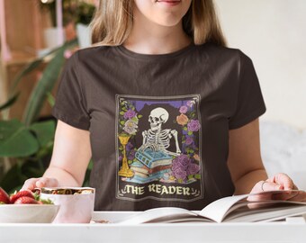 Op de afbeelding: Een bruin t-shirt met een afbeelding van een skelet dat een boek leest. De afbeelding is in een zwart kader met paarse en witte bloemen. De tekst "THE READER" staat onder de afbeelding.