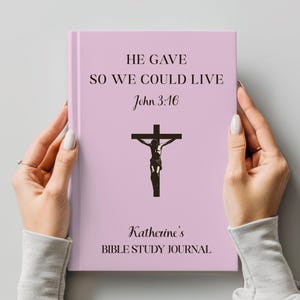 Puede incluir: Un diario de tapa dura de color rosa claro con el texto negro HE GAVE SO WE COULD LIVE John 3:16, una ilustración de un crucifijo negro y el texto personalizado Katherine's BIBLE STUDY JOURNAL impreso en la portada.