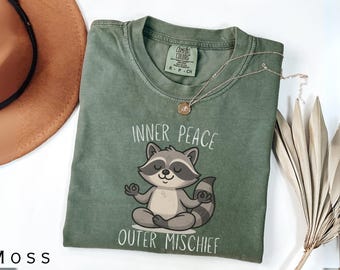 May include: A moss green t-shirt featuring a cartoon raccoon in a meditative pose. The text "Inner Peace Outer Mischief" is printed on the shirt. A gold necklace with a pendant is on the shirt. A brown hat is in the background.