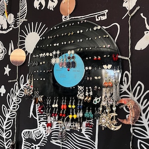 May include: A black vinyl record earring holder displaying a variety of earrings. The earrings are in various styles and colors, including studs, hoops, and dangling designs. The background is a black and white patterned fabric.