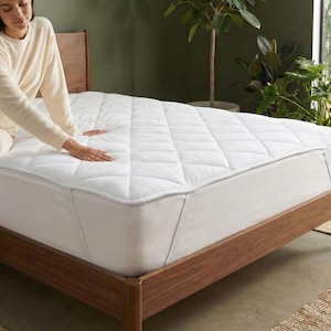 May include: A white quilted mattress topper on a wooden bed frame. The bed is in a well-lit room with plants and a neutral color palette. The topper has a diamond pattern.