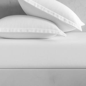 May include: Two white pillowcases with a subtle border detail rest on a white fitted sheet. The bedding is crisp and clean, suggesting a focus on comfort and simplicity. The image is well-lit, highlighting the smooth texture of the fabric.