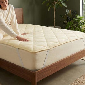 Könnte beinhalten: Ein cremefarbener Matratzenschoner mit Steppmuster liegt auf einem Bett mit Holzrahmen. Das Bett befindet sich in einem Raum mit neutraler Farbpalette und natürlichem Licht. Der Topper ist mit Riemen befestigt.