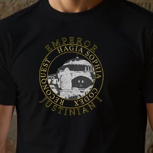 Puede incluir: Camiseta negra con un diseño circular. El diseño incluye el texto "EMPEROR HAGIA SOPHIA", "CONQUEST JUSTINIAN CODEX" y una ilustración de un edificio. La camiseta está hecha de un material suave.